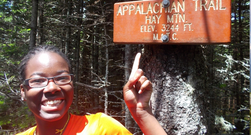 A person smiles as they point to an Appalachian trail sign posted to a tree